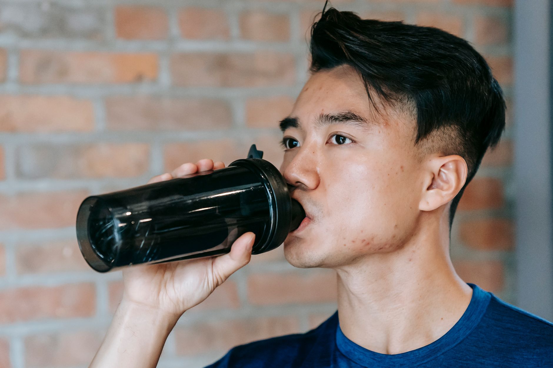 Side view of concentrated young ethnic male athlete drinking bottle of water during workout in gym