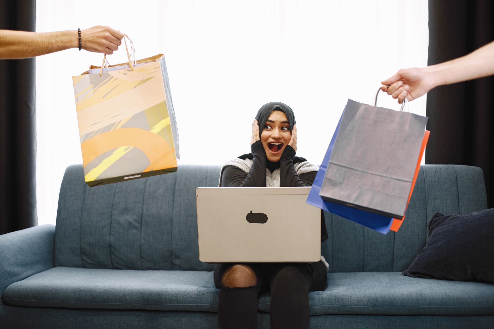 A woman in a hijab excitedly shopping online, receiving bags while sitting on a couch with a laptop.