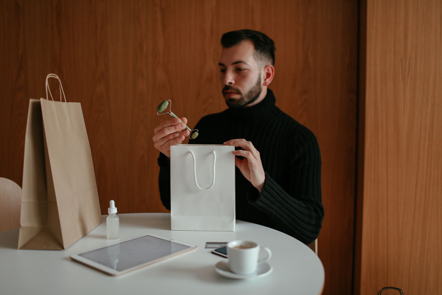 Confident young bearded metrosexual male in warm sweater pulling out jade roller face massager from white paper bag after making online order with credit card and tablet