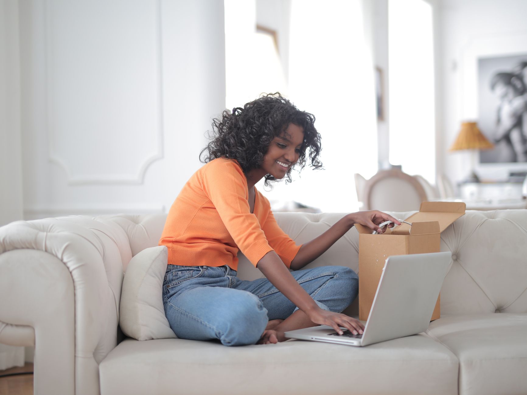 Side view of African American lady in casual wear sitting with crossed legs on sofa while opening carton box and using laptop