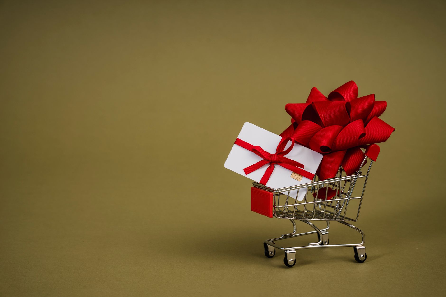 Mini shopping cart containing a gift card adorned with a red bow on a neutral background, ideal for retail concepts.