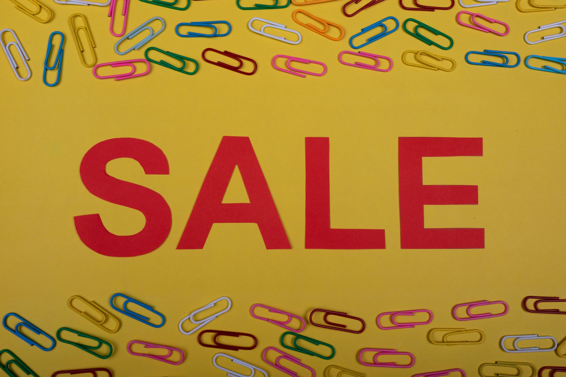 Bright sale sign surrounded by colorful paper clips on a vibrant yellow background.