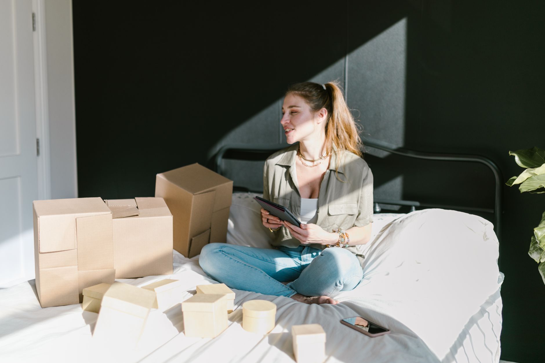 Woman sitting on bed with boxes and tablet, running an online business.