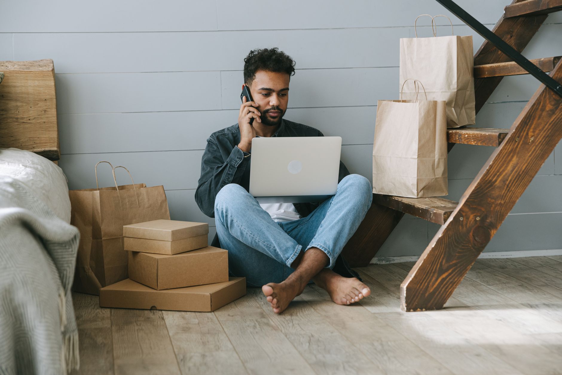 Adult man working remotely on laptop, surrounded by shopping bags and boxes indoors.