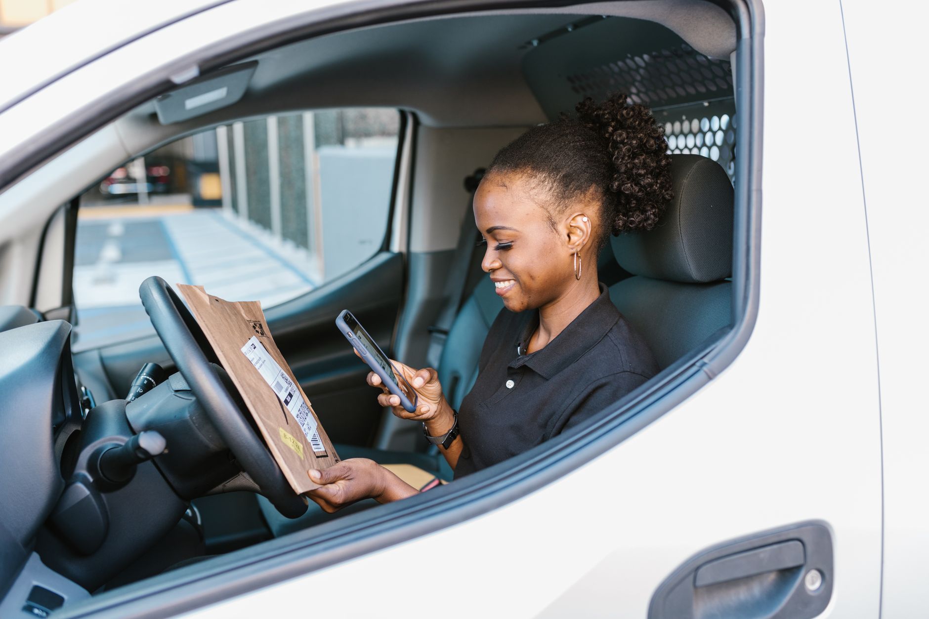 Female courier using smartphone while holding a parcel inside a car. Efficient delivery service in action.