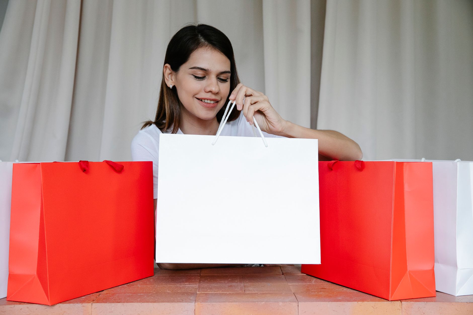 Excited young female shopaholic with long dark hair smiling while putting paper bags on brick surface after successful shopping