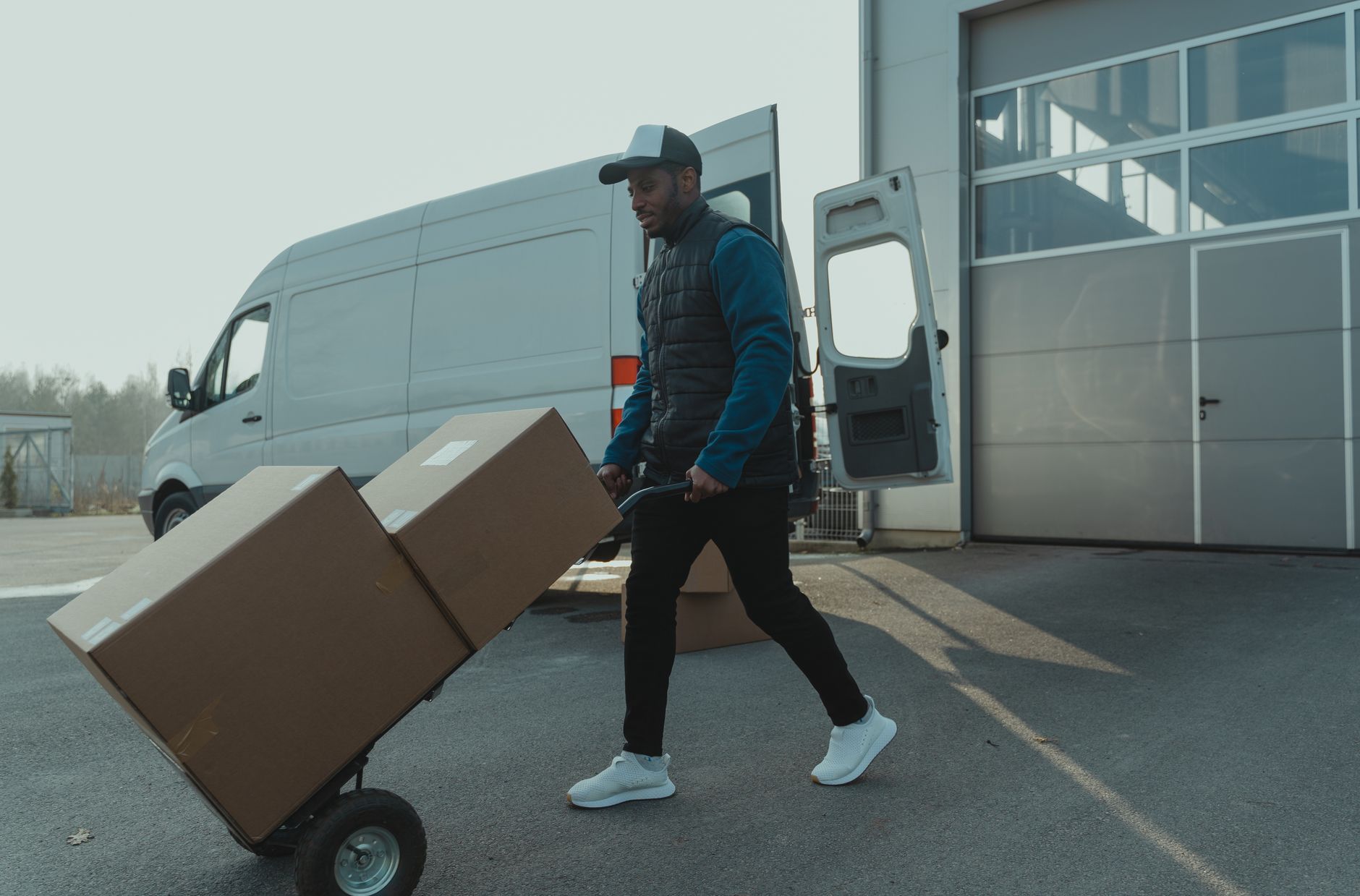 Courier pushing a trolley with boxes near a van at a warehouse, showcasing delivery services.