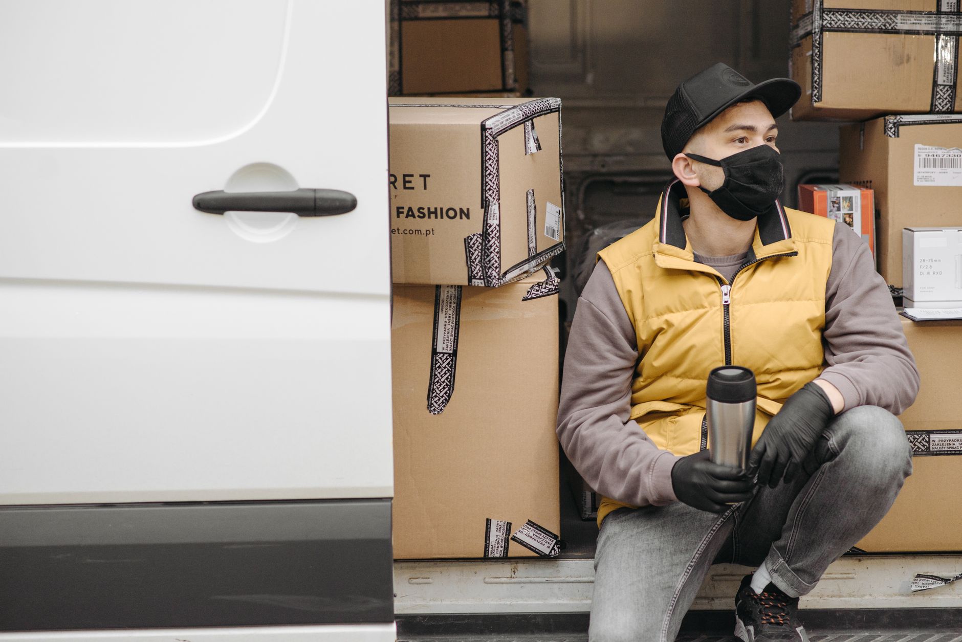 A courier wearing a face mask sits by a delivery van with parcels, ready for shipment.