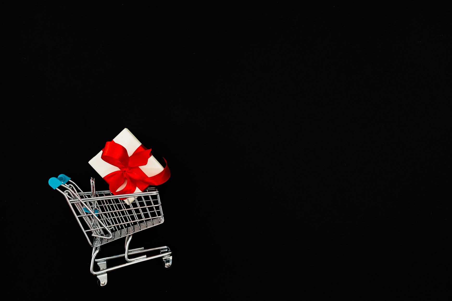 A shopping cart carrying a gift box with a red ribbon, set against a dark background.