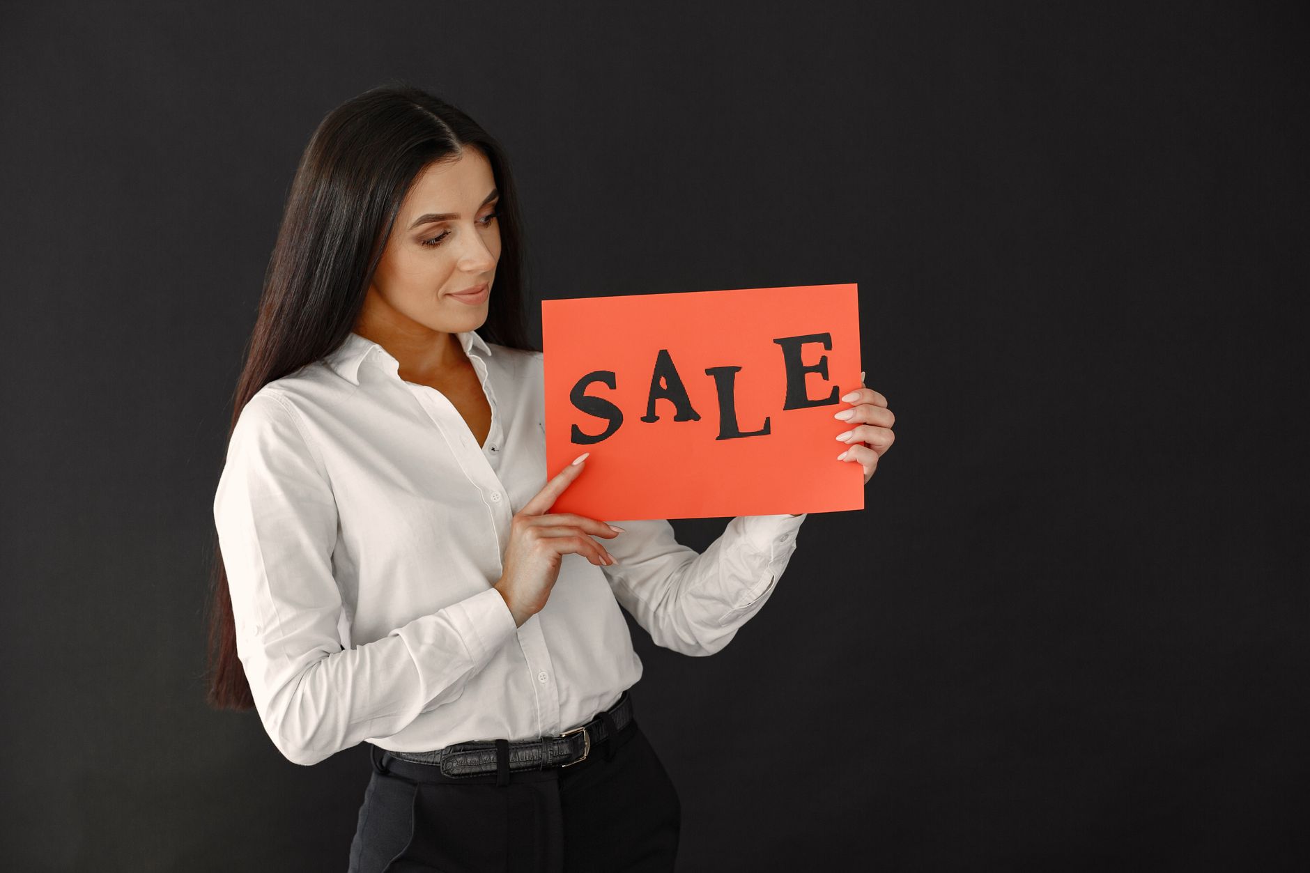 A woman in a white shirt holds a red sale sign against a dark background in a studio setting.
