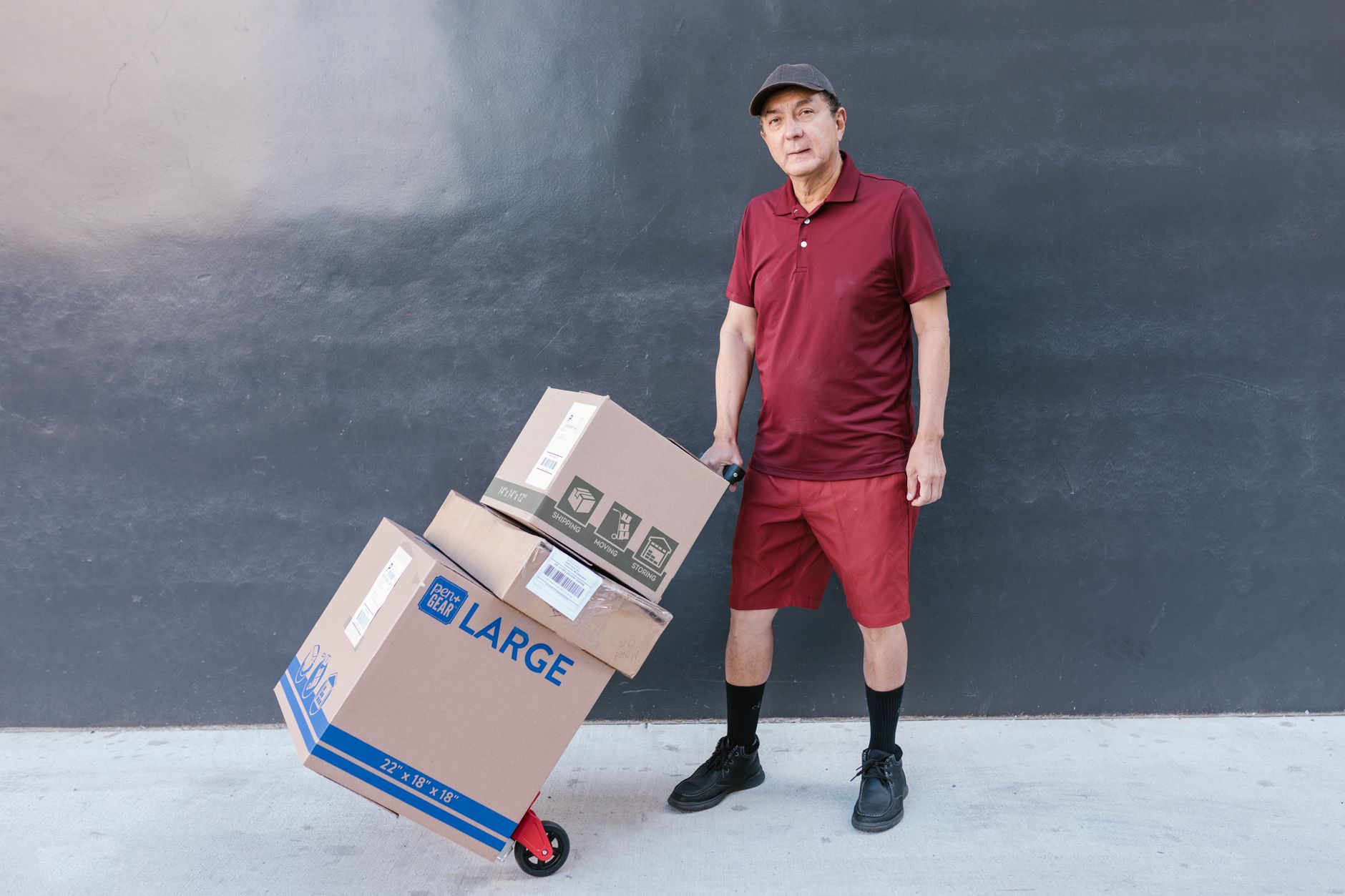 Delivery worker in red uniform with boxes on hand truck, standing against a gray wall.