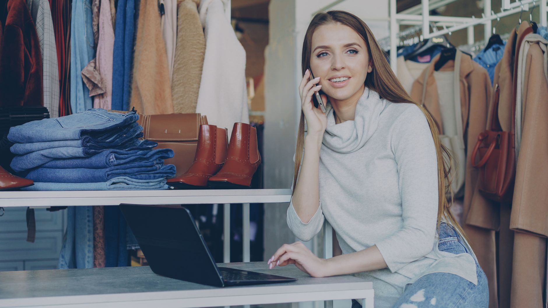 Woman in boutique talks on phone with laptop, surrounded by clothes.