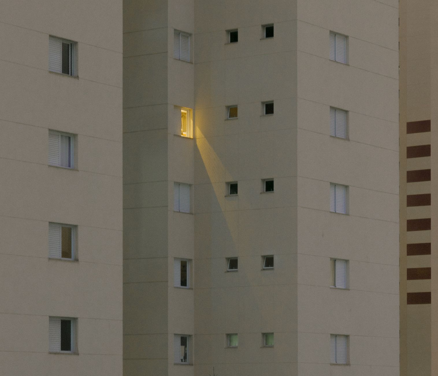 A modern apartment building facade at night with one lit window, creating a striking focal point.