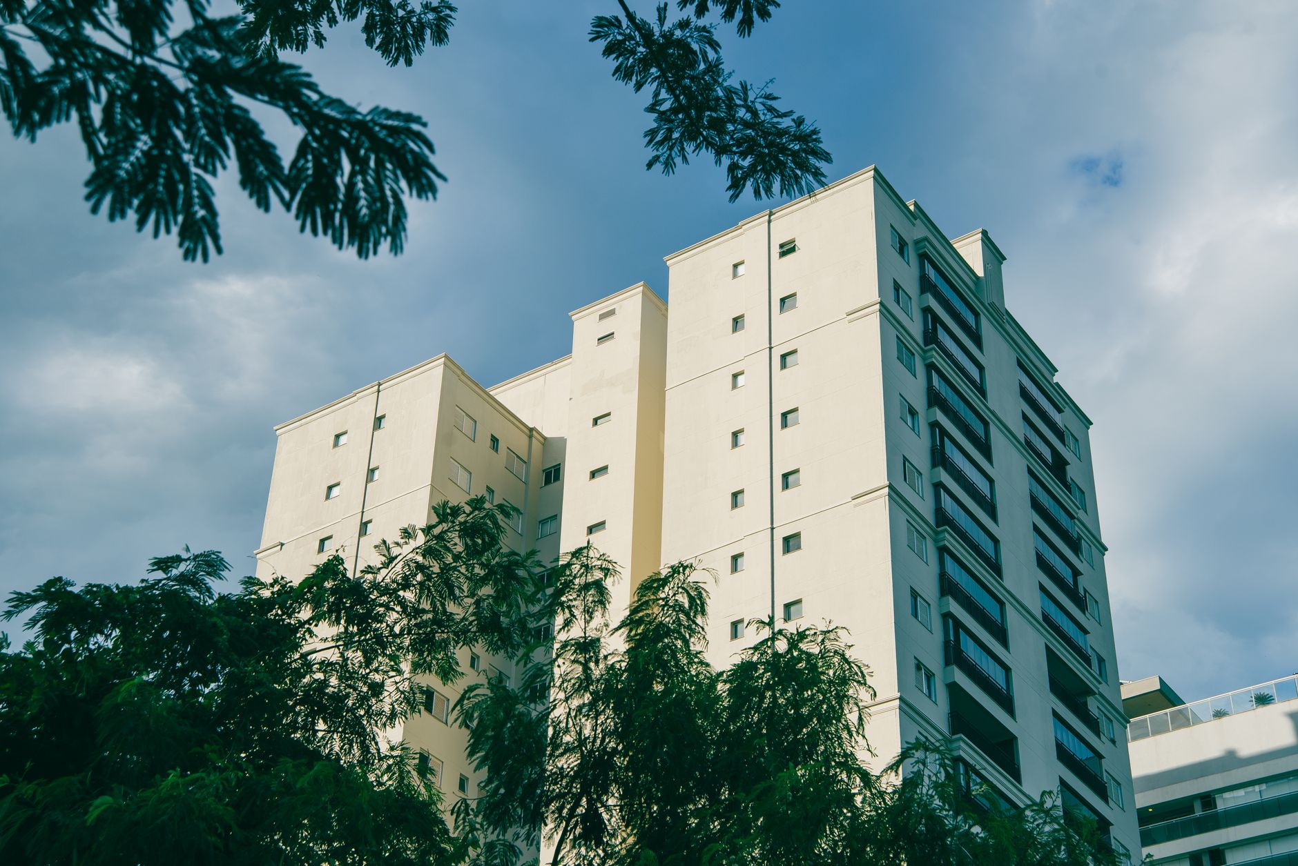 A tall residential building with green trees against a cloudy blue sky.