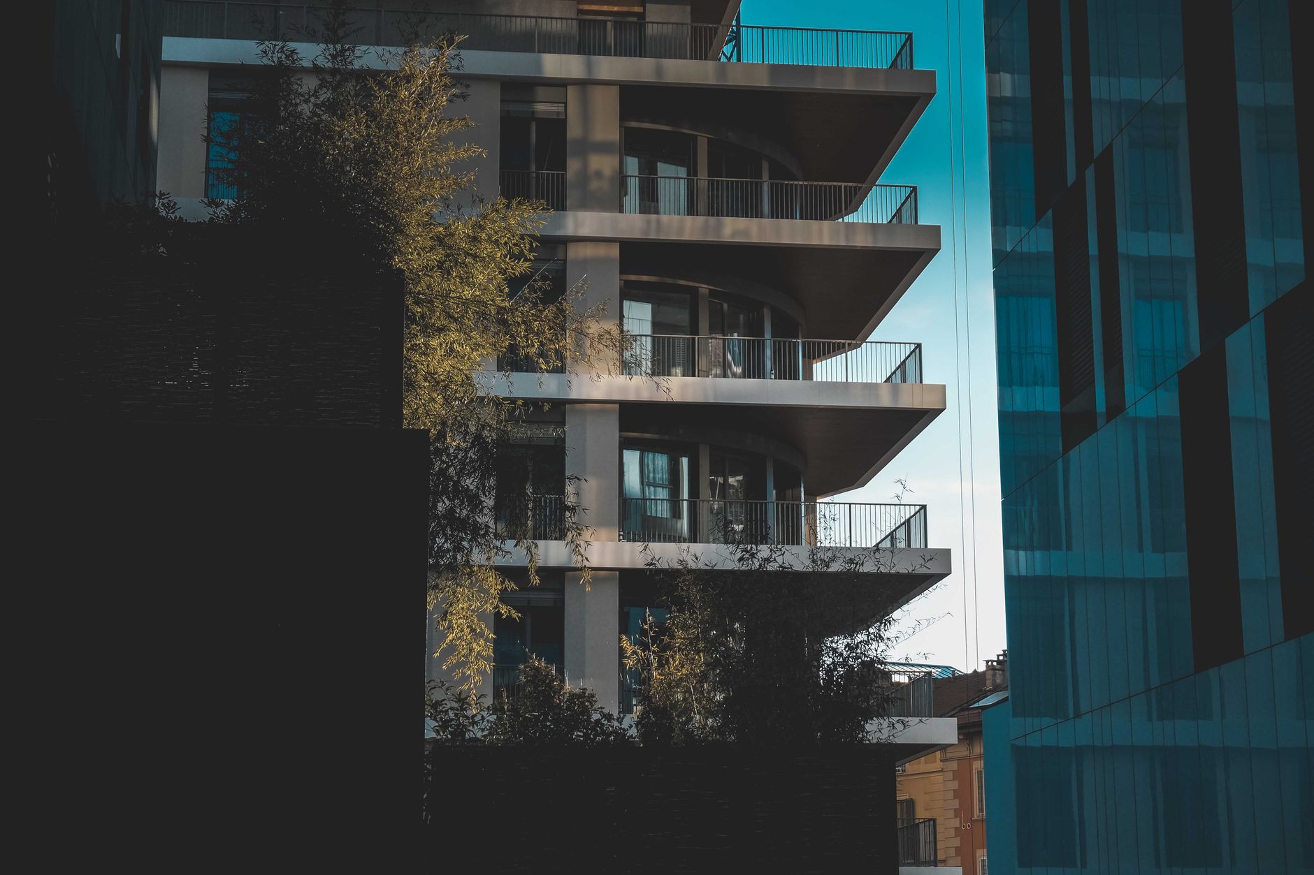 Contemporary residential multistory house with balconies and metal fences located on street with tree and shadow in city against blue sky