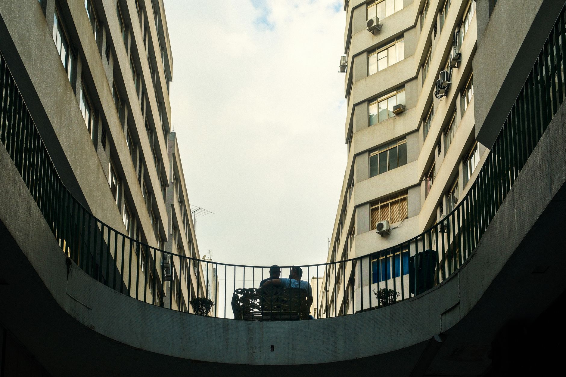 Silhouettes of two people chatting on a balcony between tall apartment buildings in a city.