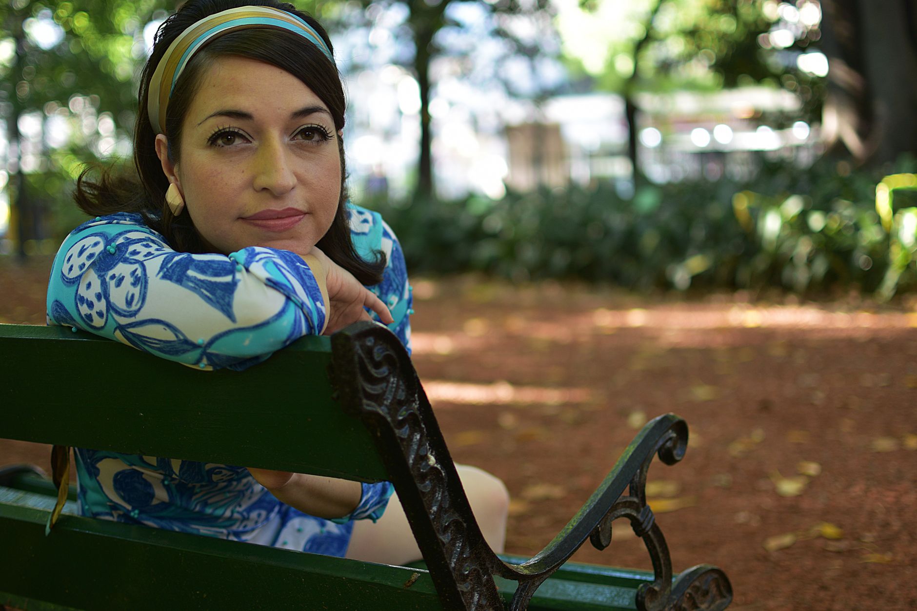 A woman in a blue floral dress relaxes on a green bench in a sunny park setting.