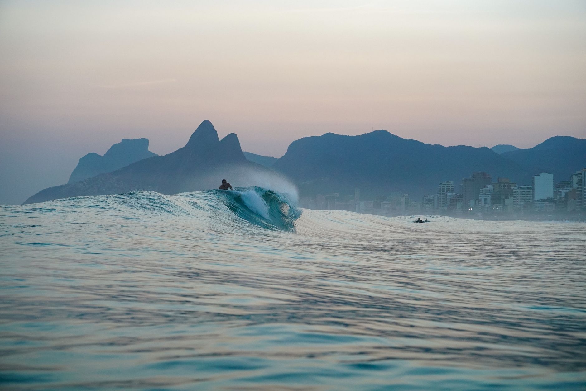 A surfer enjoys the serene waves of Ipanema Beach at sunset, with Two Brothers Mountain in the background.
