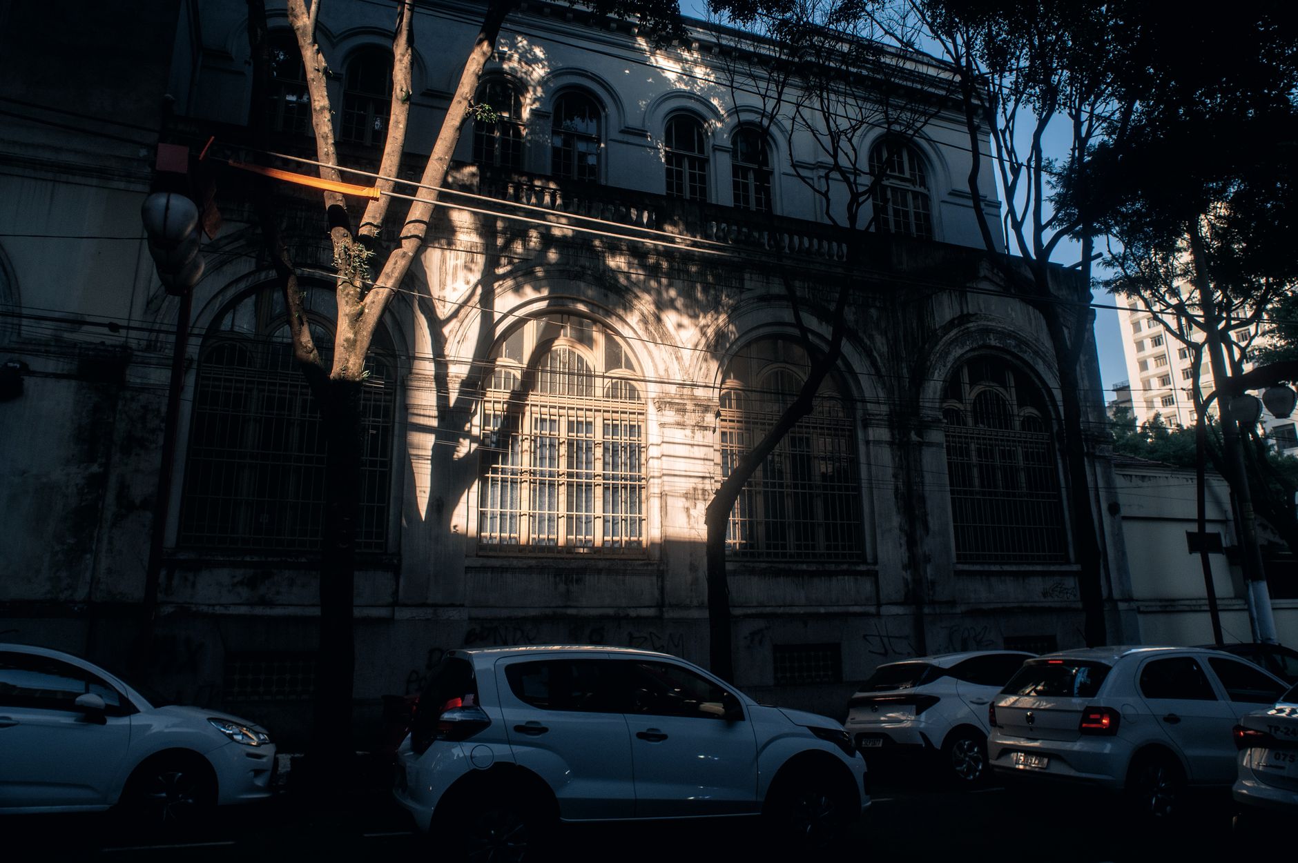 A historic building facade with cars parked under tree shadows in evening light.