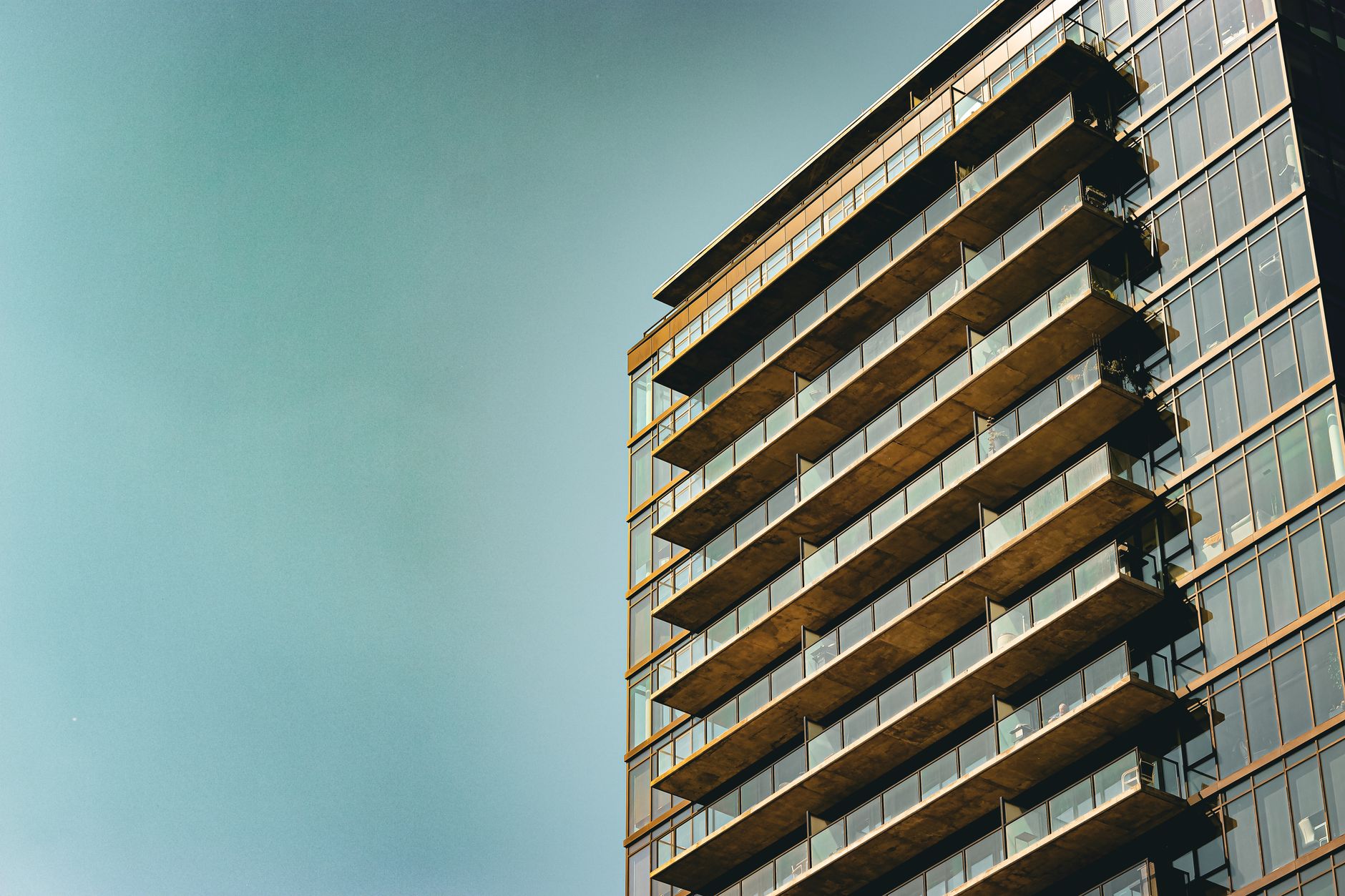 Sleek modern apartment building with glass facade and balconies, set against a clear blue sky.