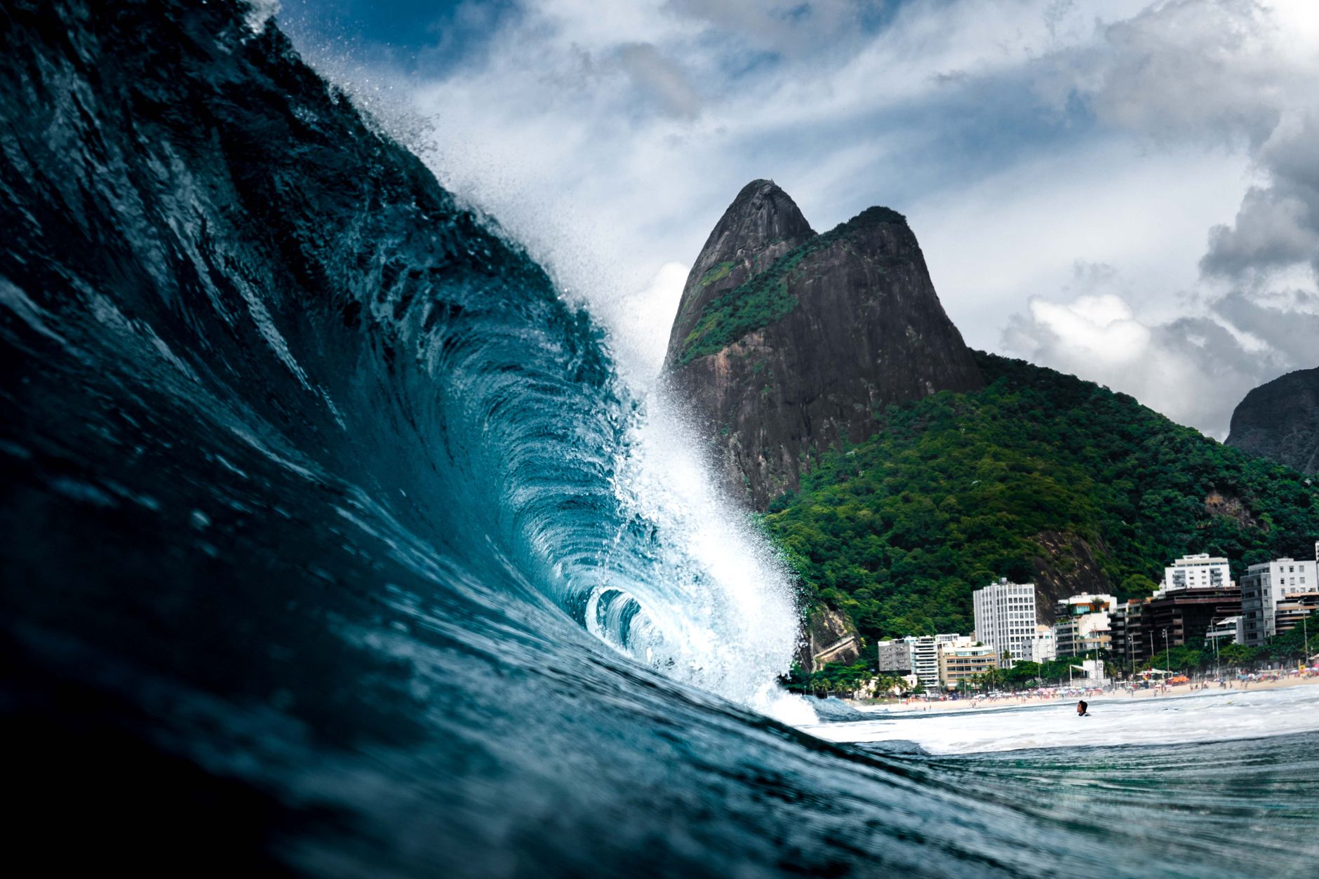 Powerful wave crashing near Brazil's iconic coastline with lush green mountains and city view.