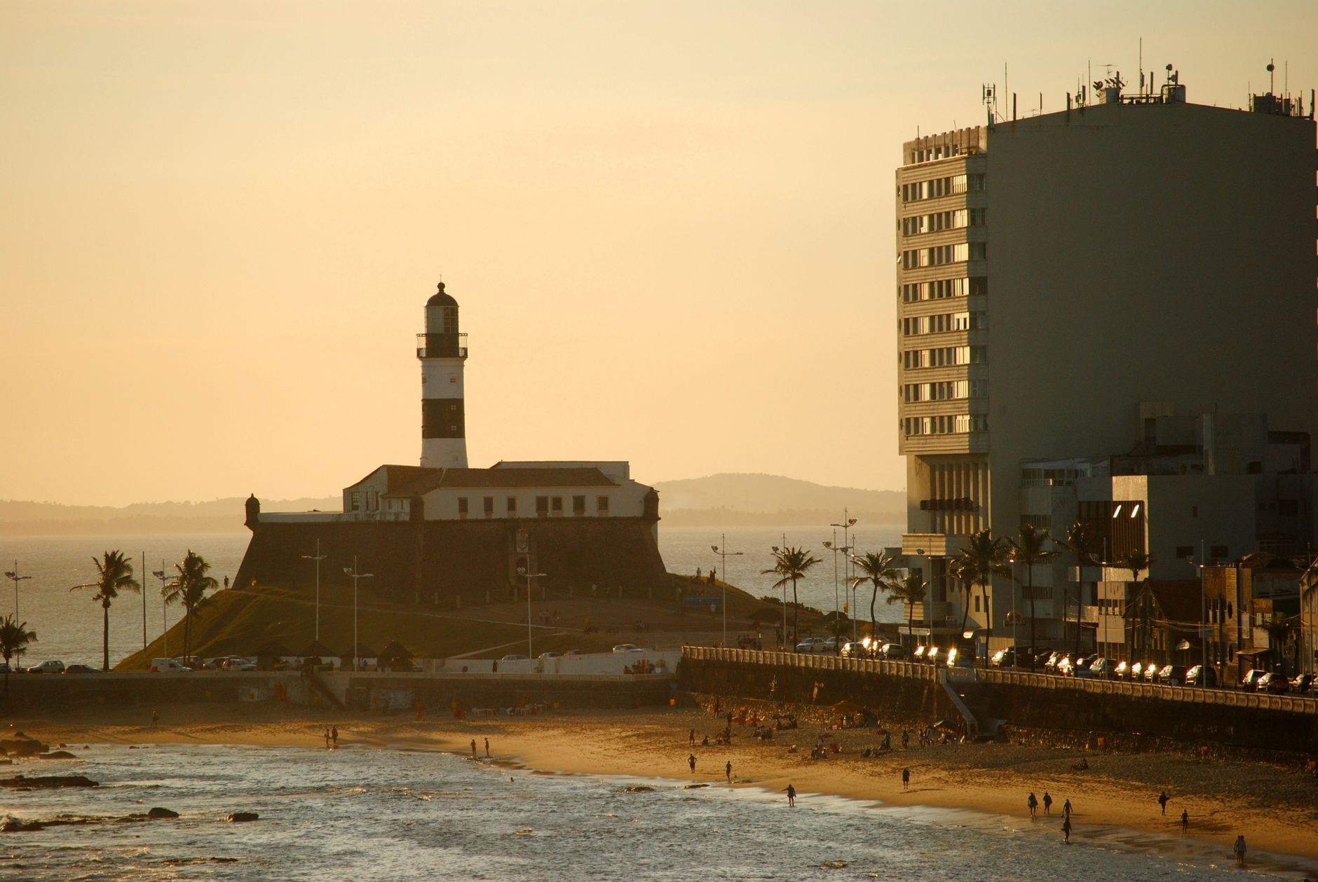 Stunning view of the Farol da Barra lighthouse in Salvador, Brazil during sunset, with tourists on the beach.