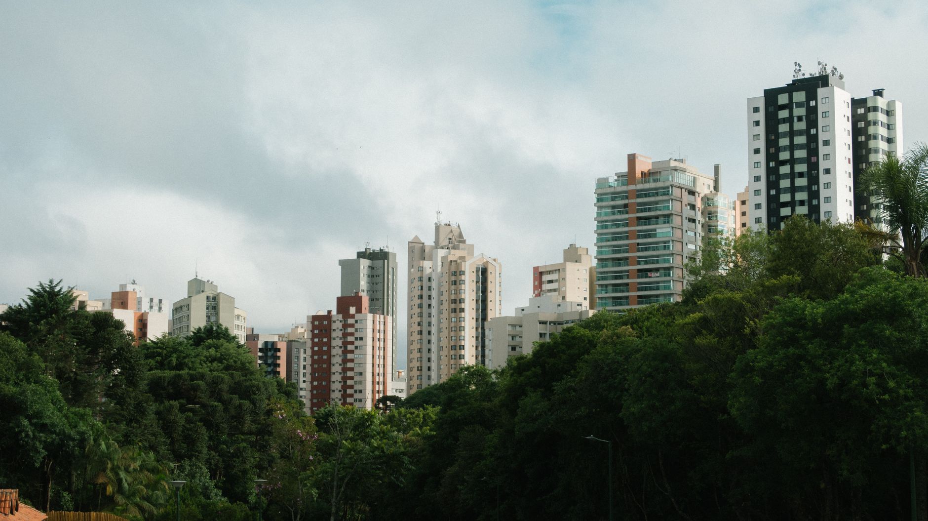 Cityscape featuring a cluster of tall buildings behind lush greenery under a cloudy sky.