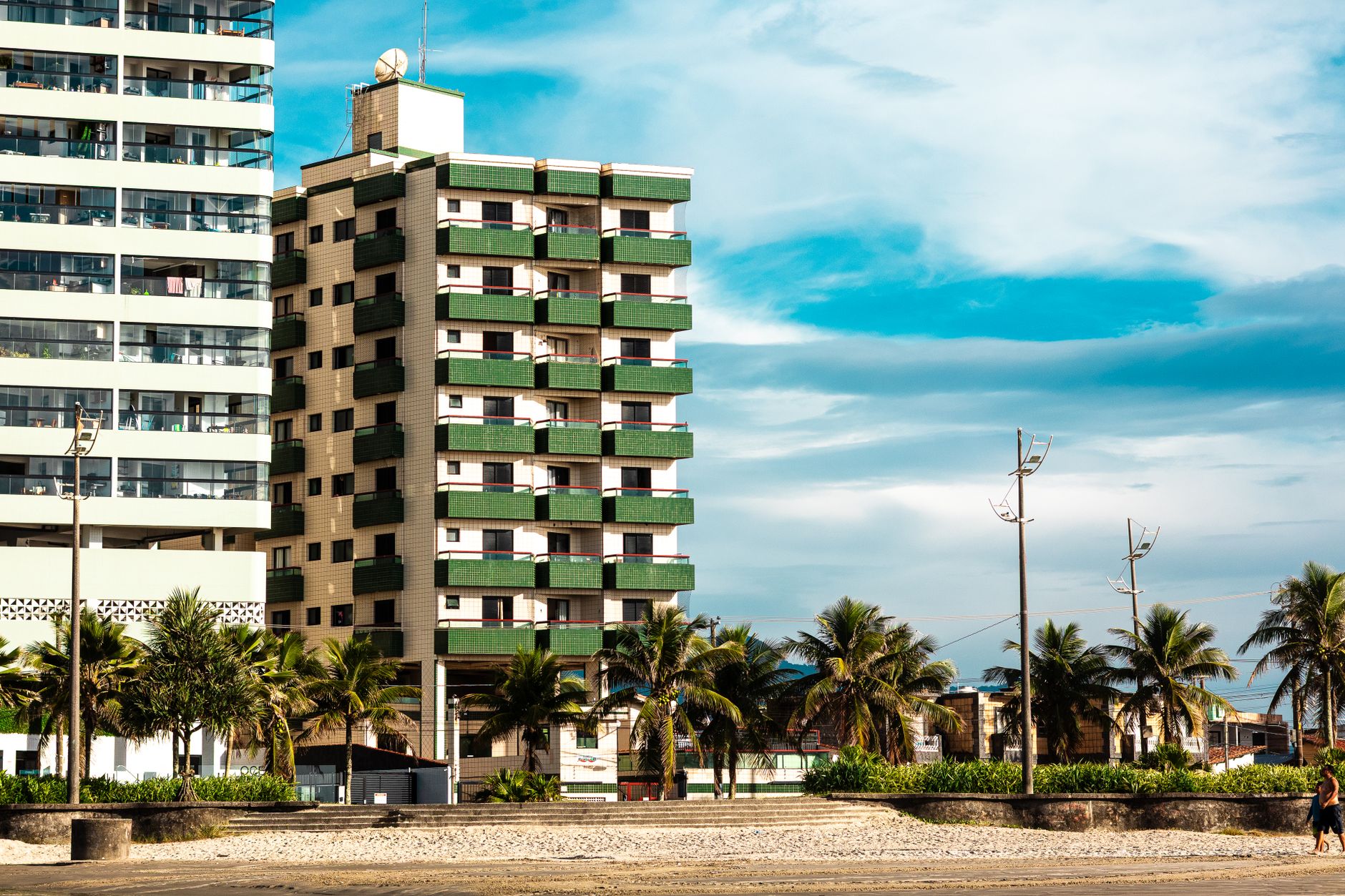 A modern apartment building next to a tropical beach with palm trees under a clear blue sky.