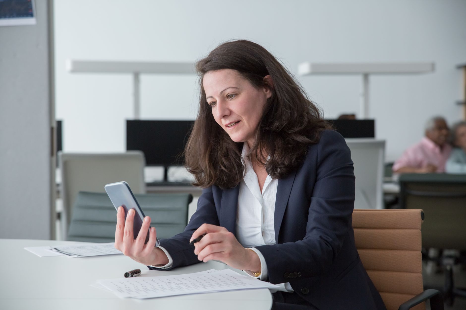 Professional woman in an office setting using a smartphone and writing notes at a desk.