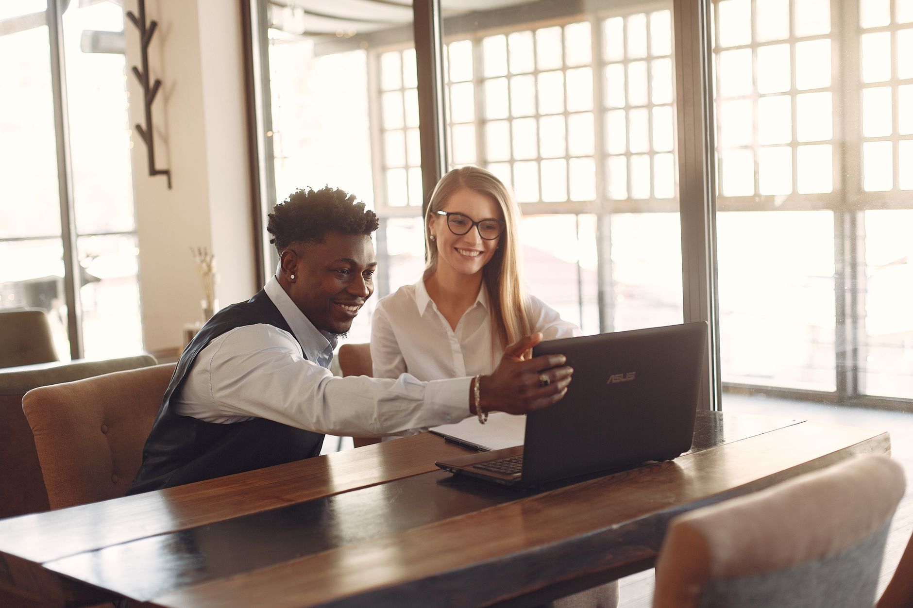 Cheerful young multiethnic business partners in formal clothes sitting together at wooden table with laptop and discussing results of work in modern workspace
