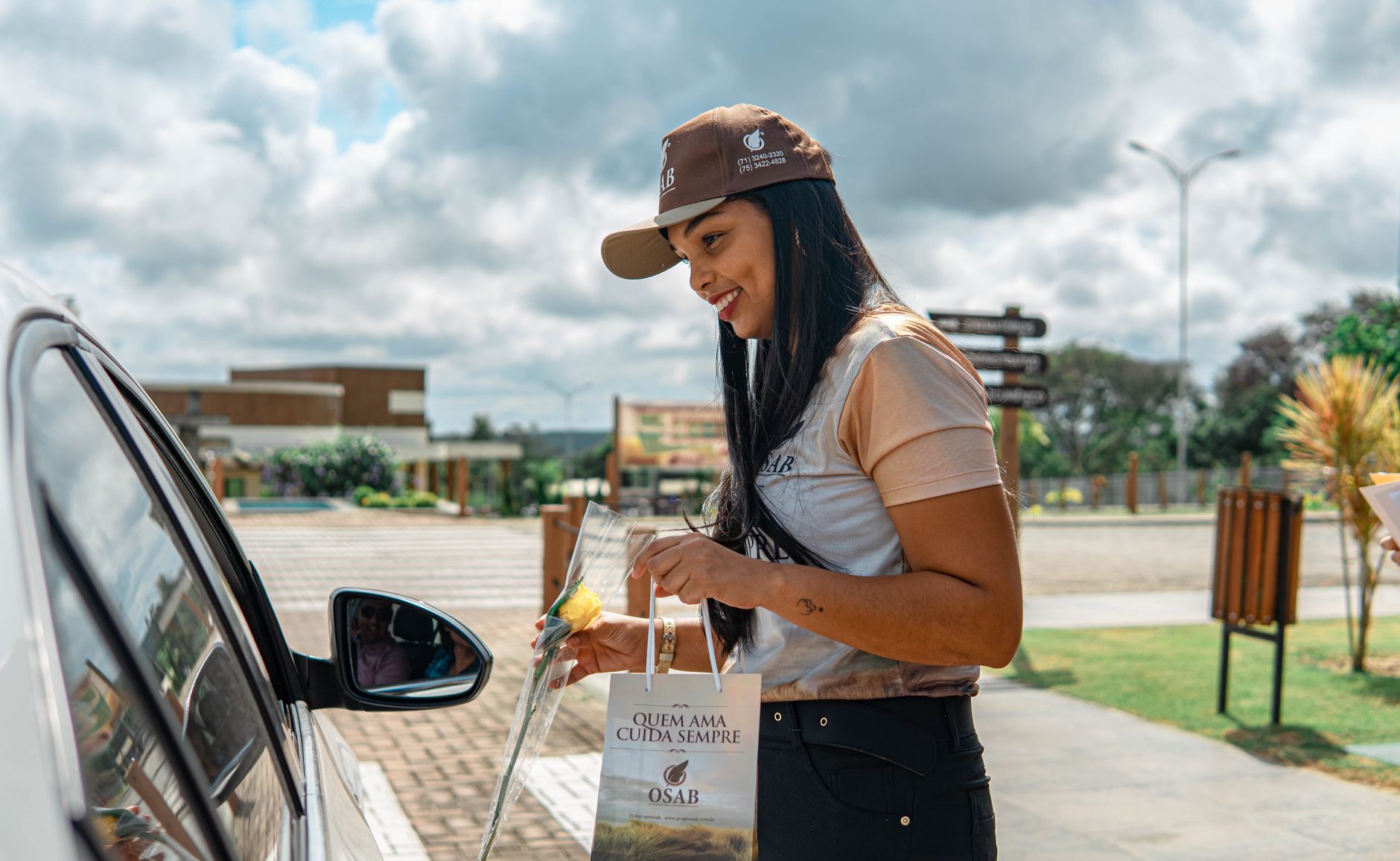 A smiling woman handing a promotional gift to a driver through a car window outdoors.