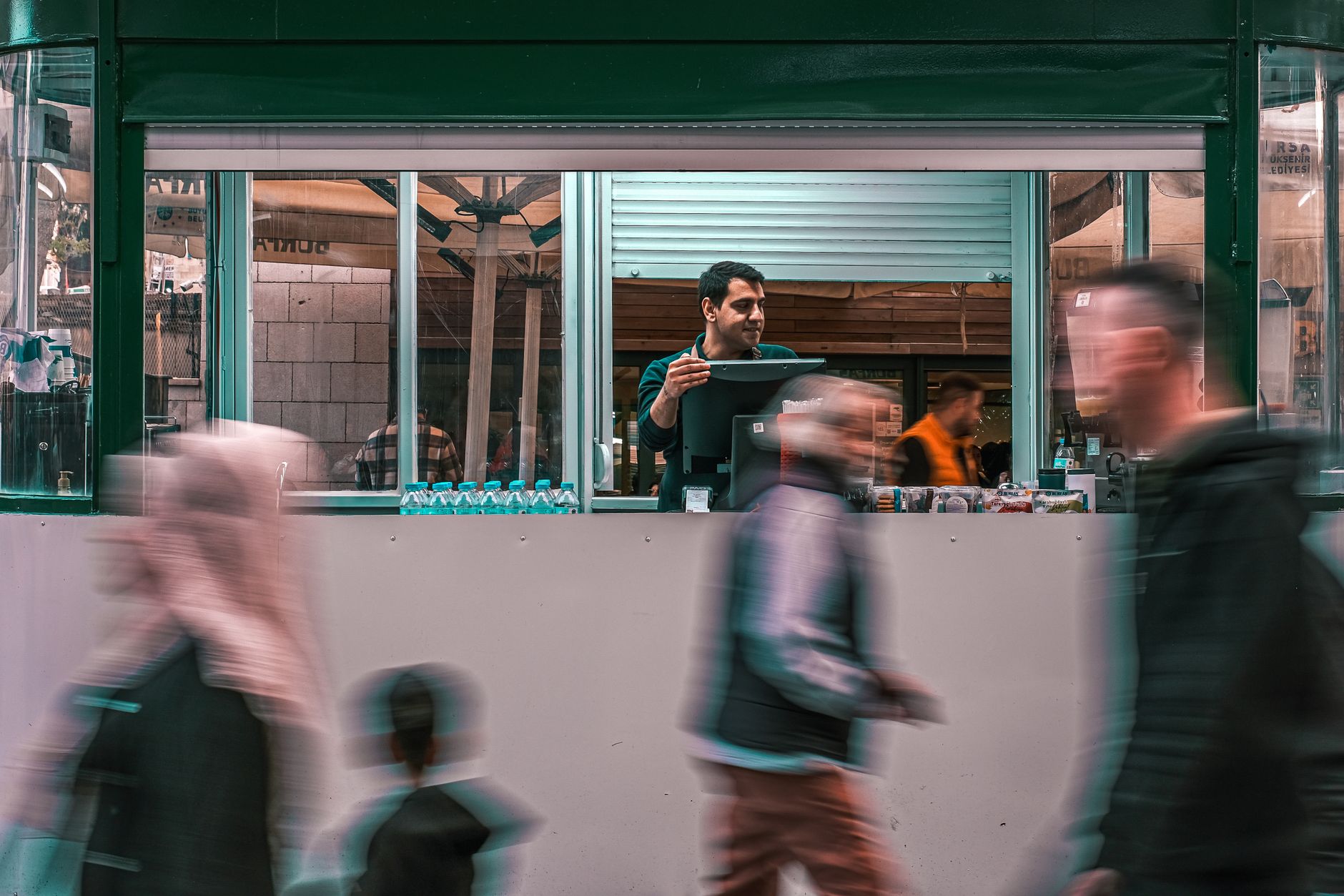 A street vendor serving customers through a kiosk window while people walk by in a busy market setting.