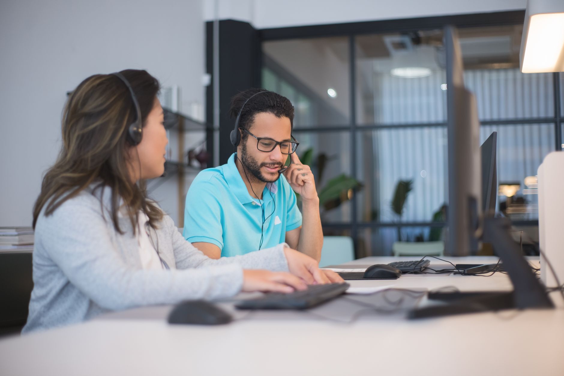 Two call center employees working together in a modern office setting with headsets and computers.