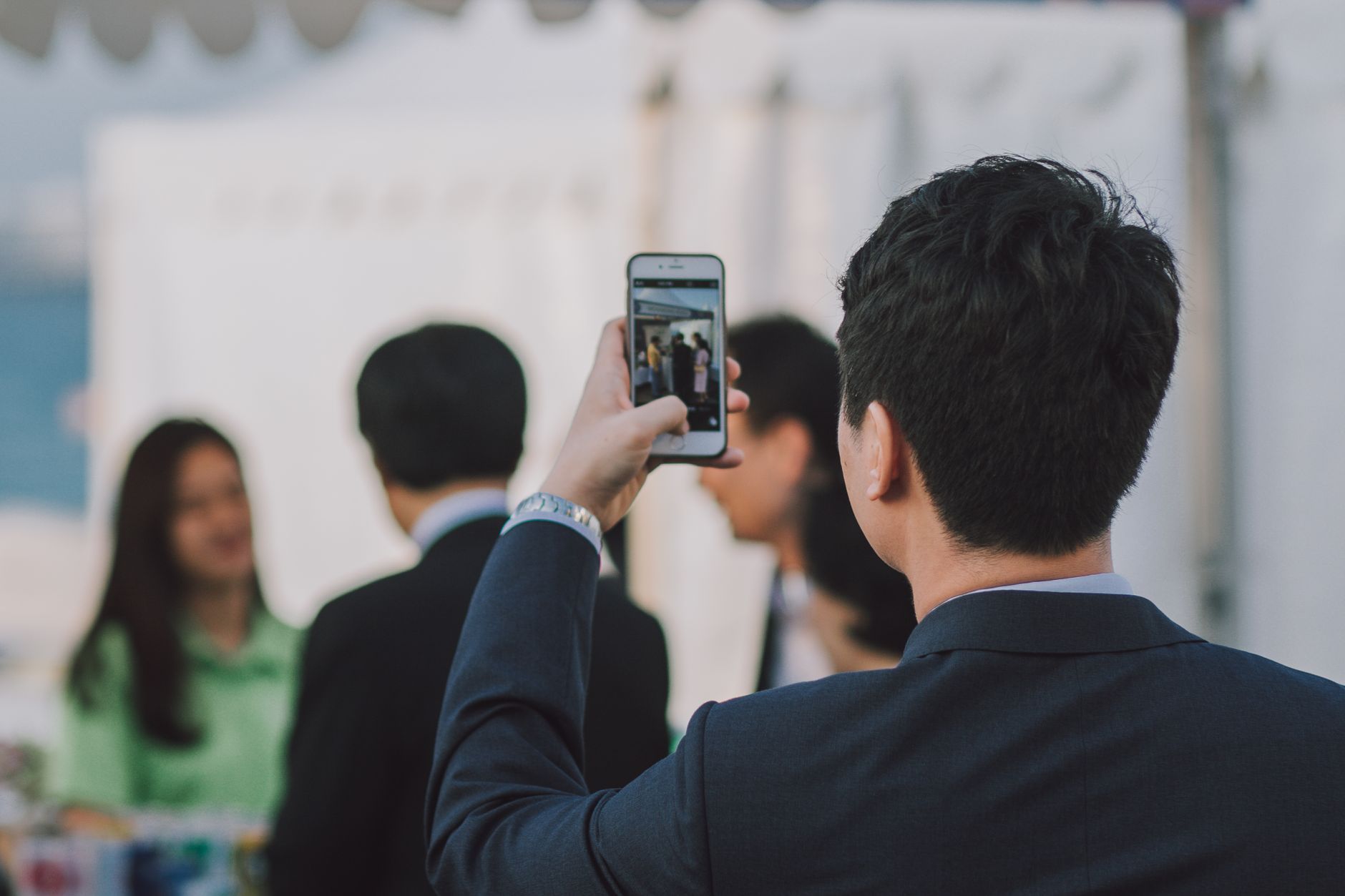 Man in suit photographing colleagues with smartphone during business event outdoors.