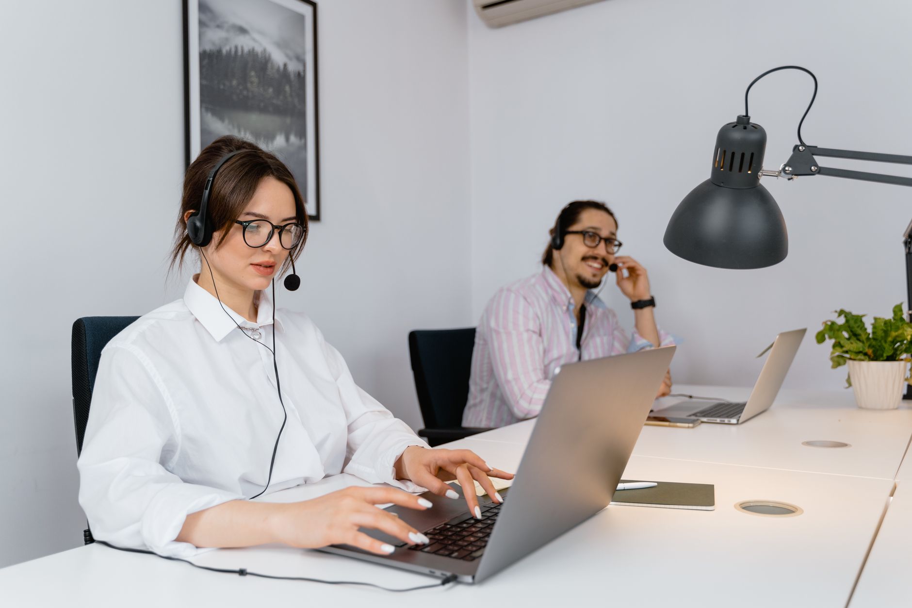 Two call center employees working with headsets and laptops in a modern office setting.