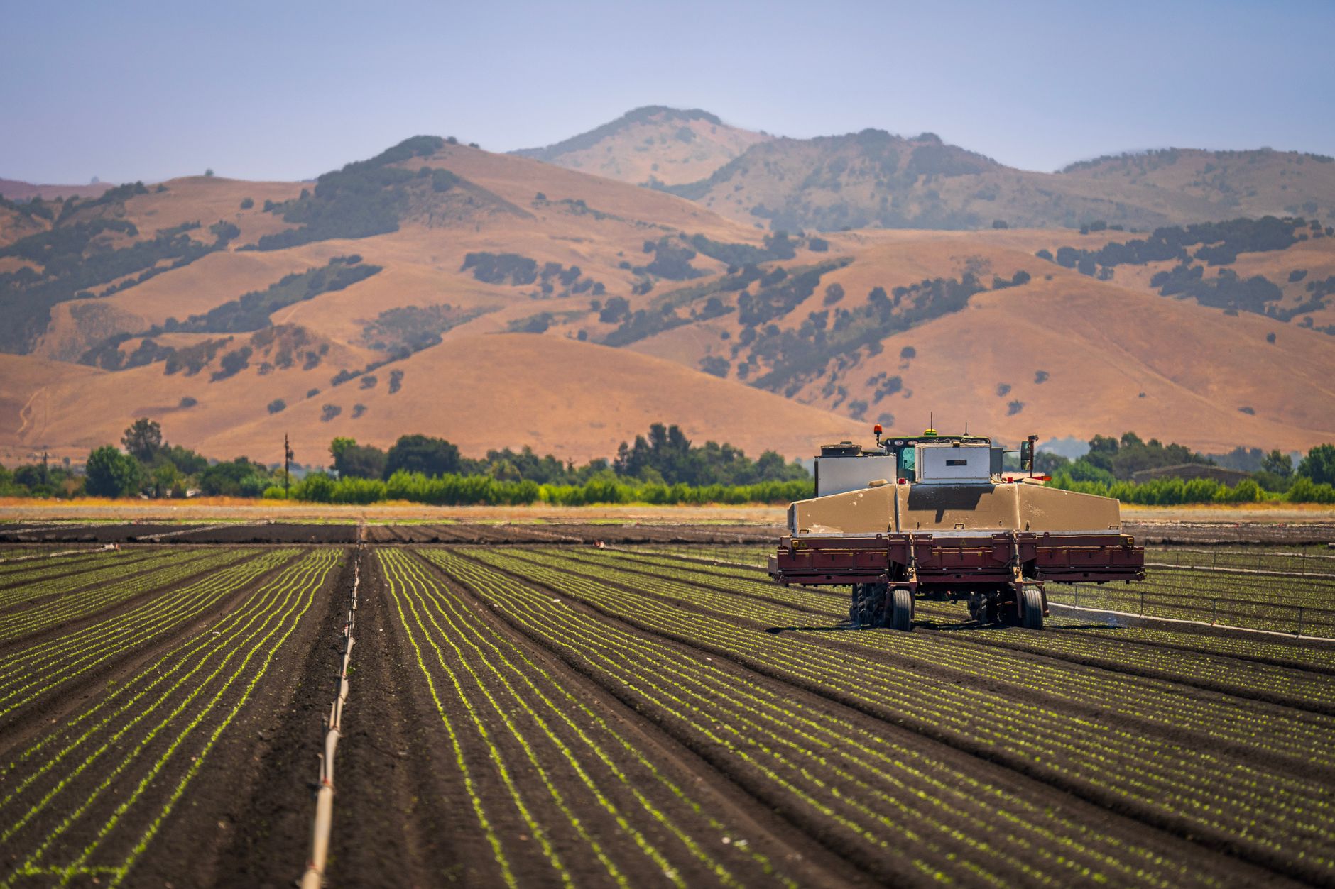 A farming machine operates on a sprawling agricultural field with hilly countryside views.