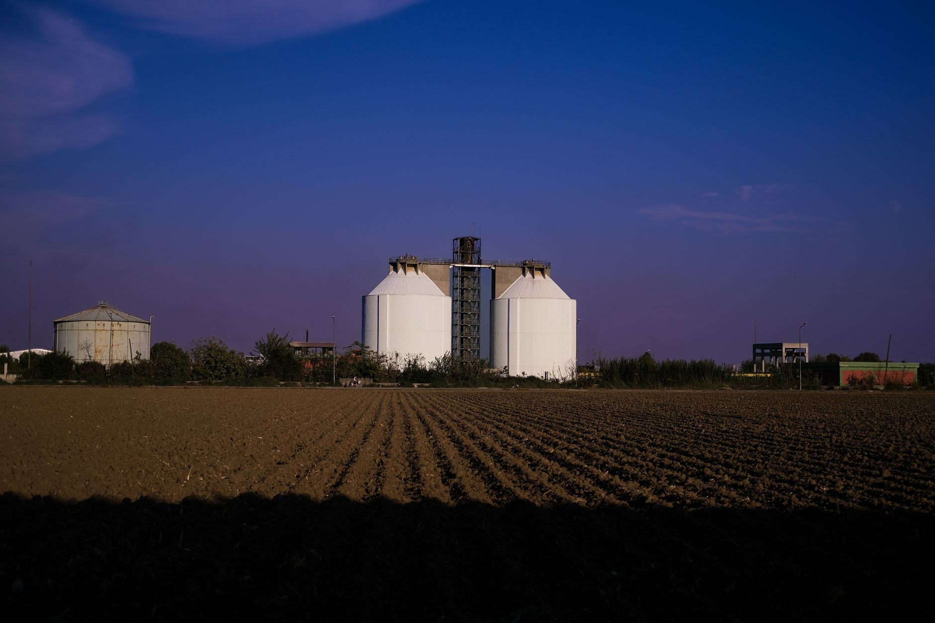 White industrial silos stand in a field during twilight in Adana, Türkiye, creating a striking silhouette against the evening sky.