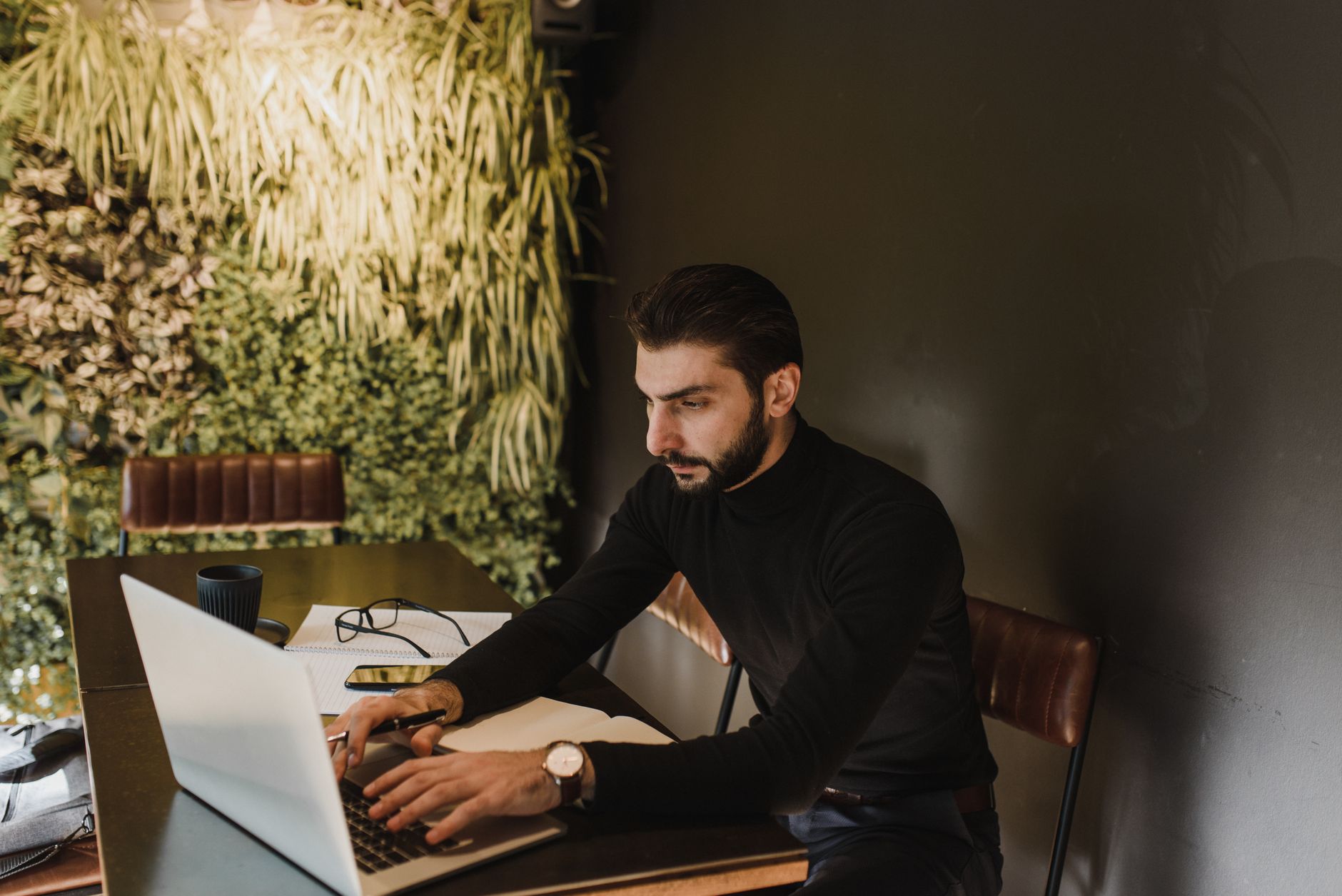 Businessman working on laptop in a stylish indoors office with greenery wall and sleek furniture.