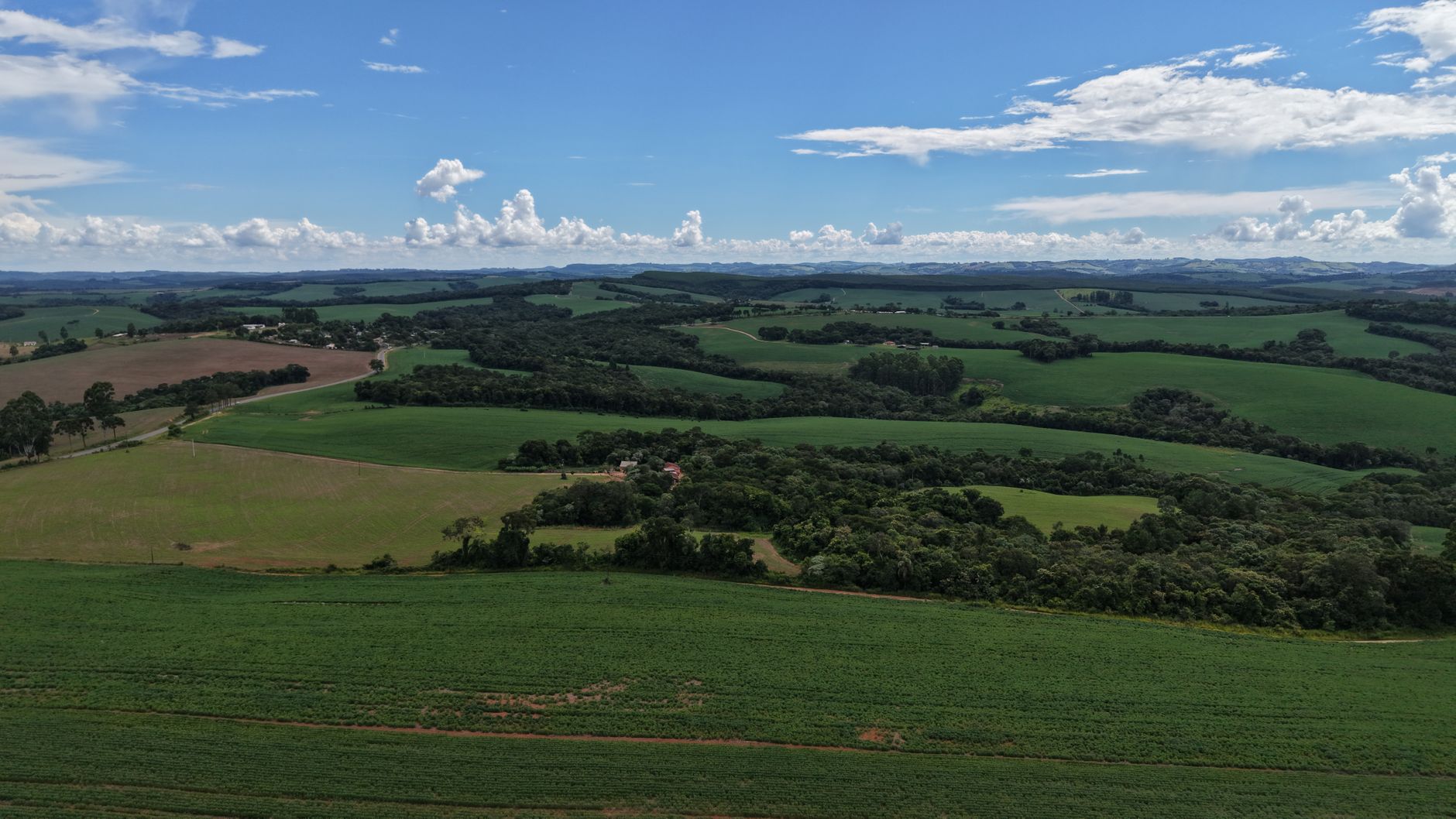 Scenic aerial view of green rolling fields in Brazil beneath a clear blue sky with scattered clouds.