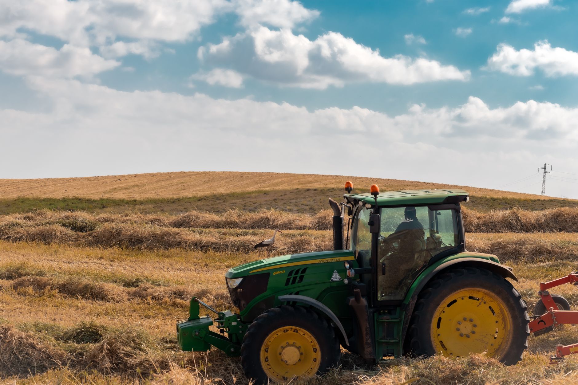 A green tractor plows through a rural field under a bright blue sky with clouds.
