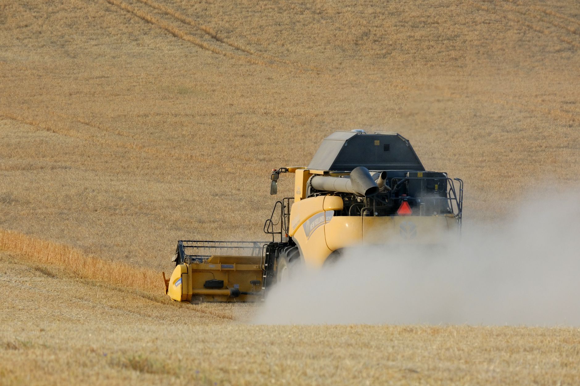 A yellow harvester tractor plows through a dusty wheat field under the summer sun.