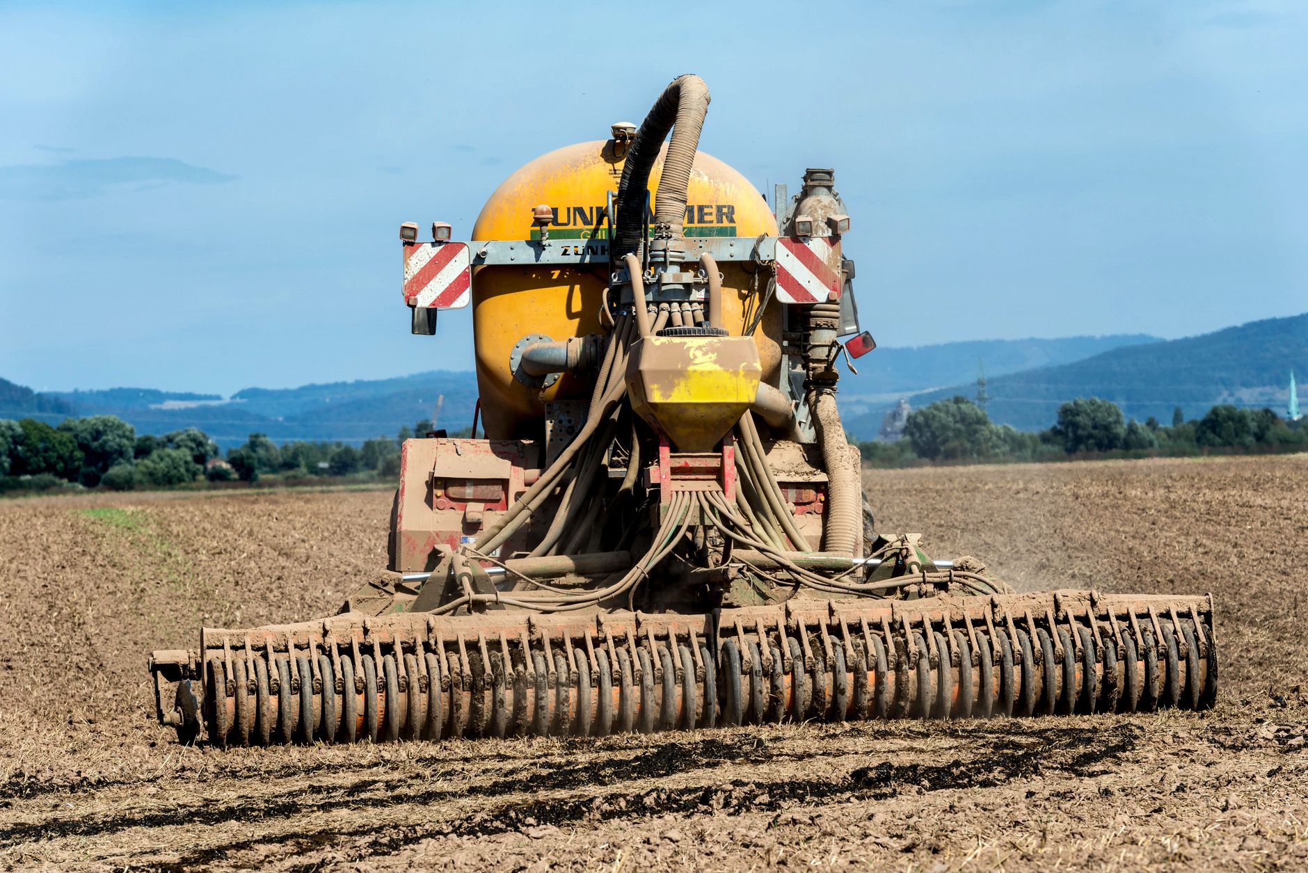 A rear view of farm machinery fertilizing a field in the countryside under a clear blue sky.
