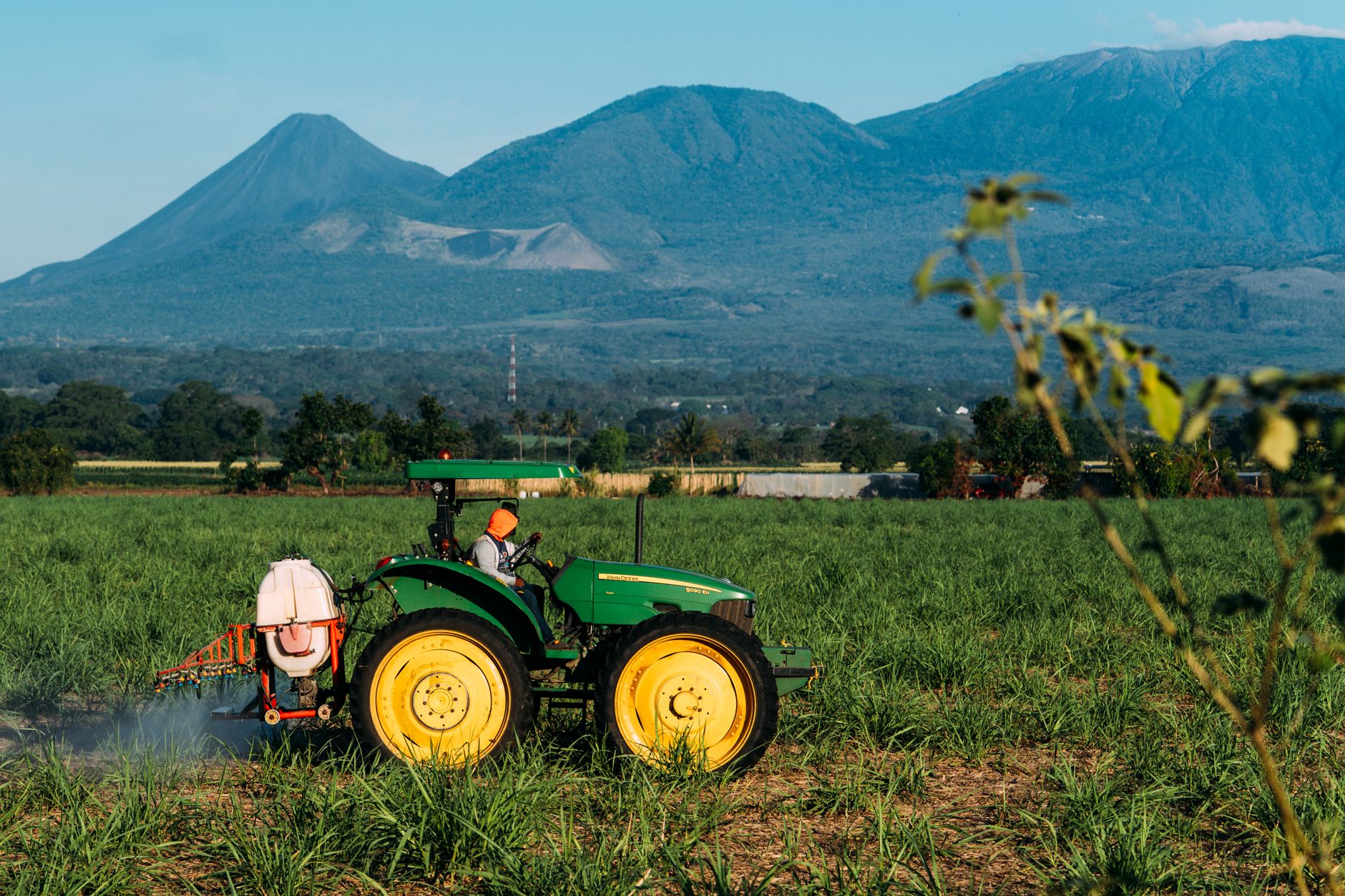 A tractor sprays crops in a lush field with majestic mountains in the background under a clear blue sky.
