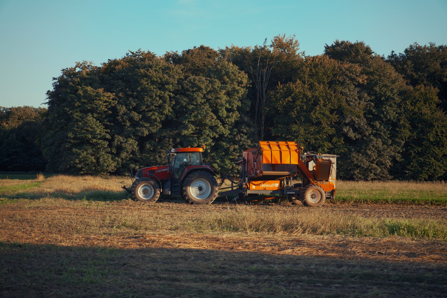 Tractor and harvester working in a field next to a forest during autumn.