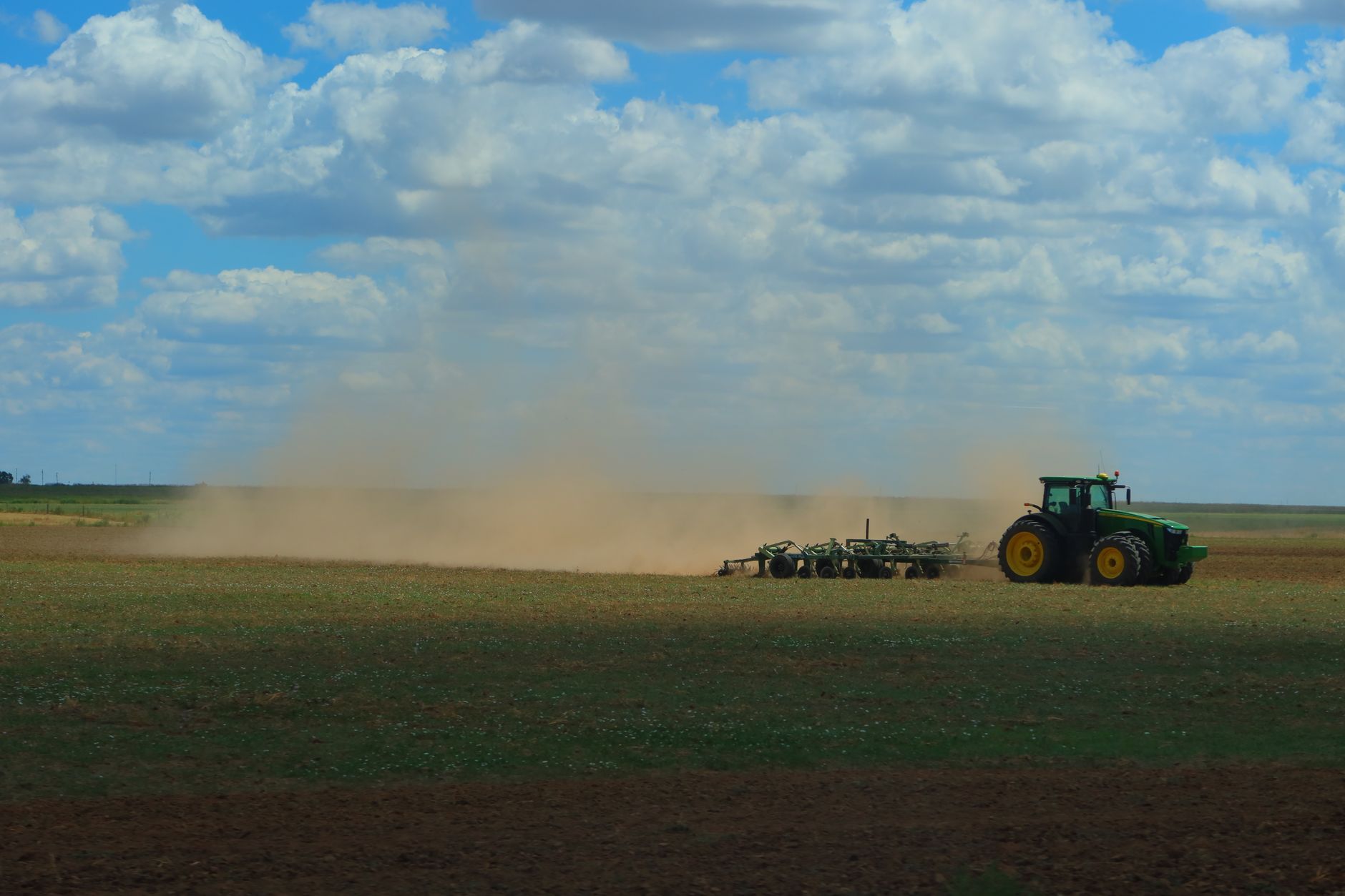 A tractor working in a dusty field under a bright blue sky with clouds.