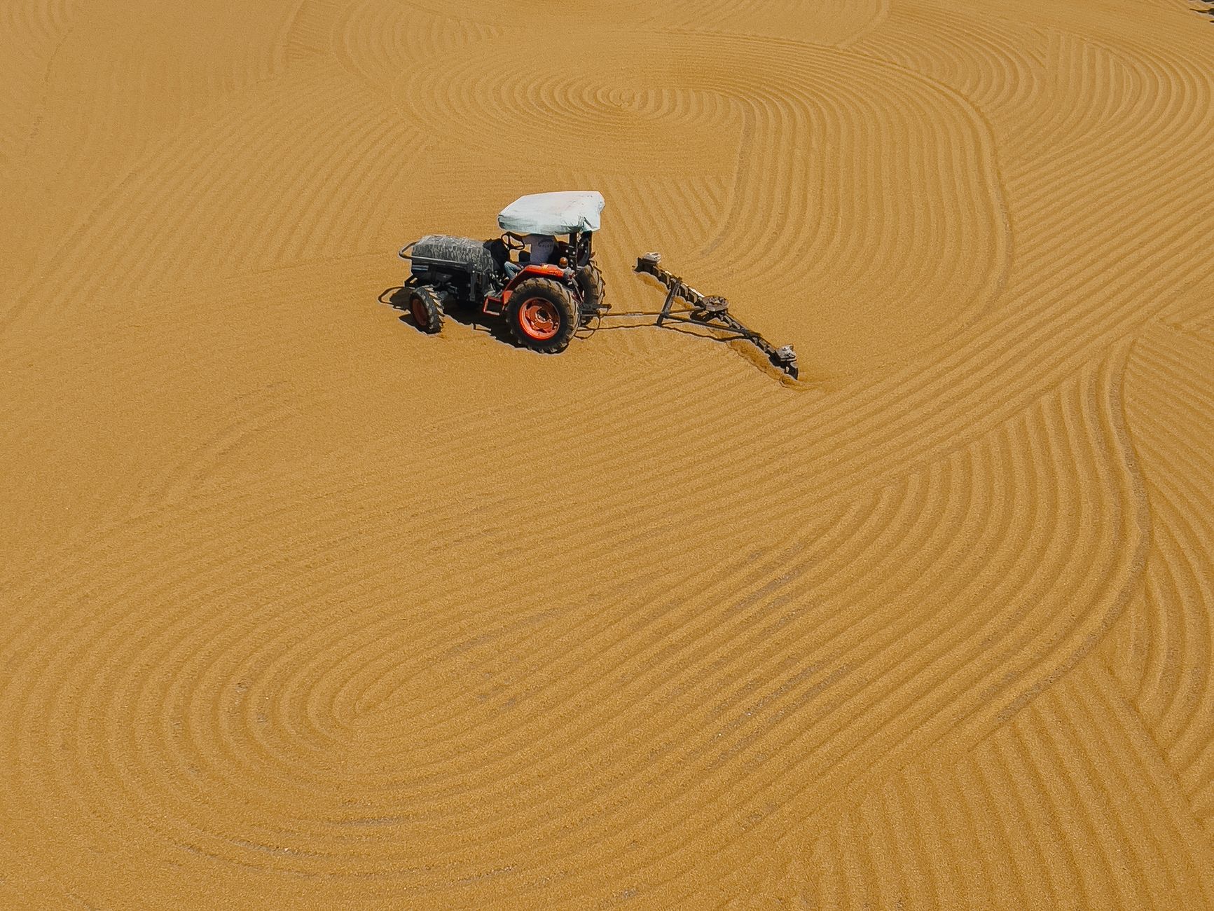 A tractor creates intricate patterns in a vast sandy field, captured from above.