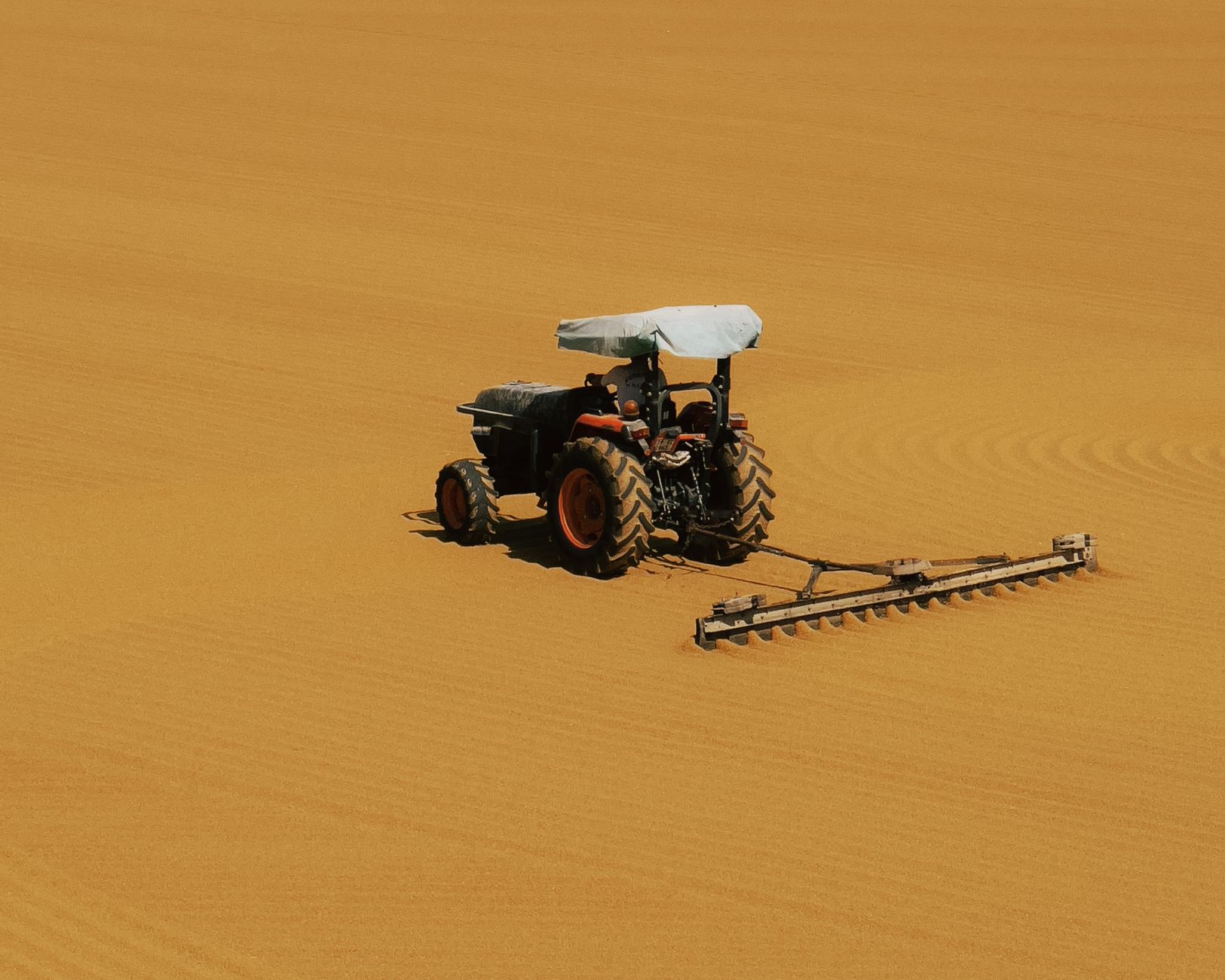 A solitary tractor works on endless sand dunes, emphasizing isolation and scale.