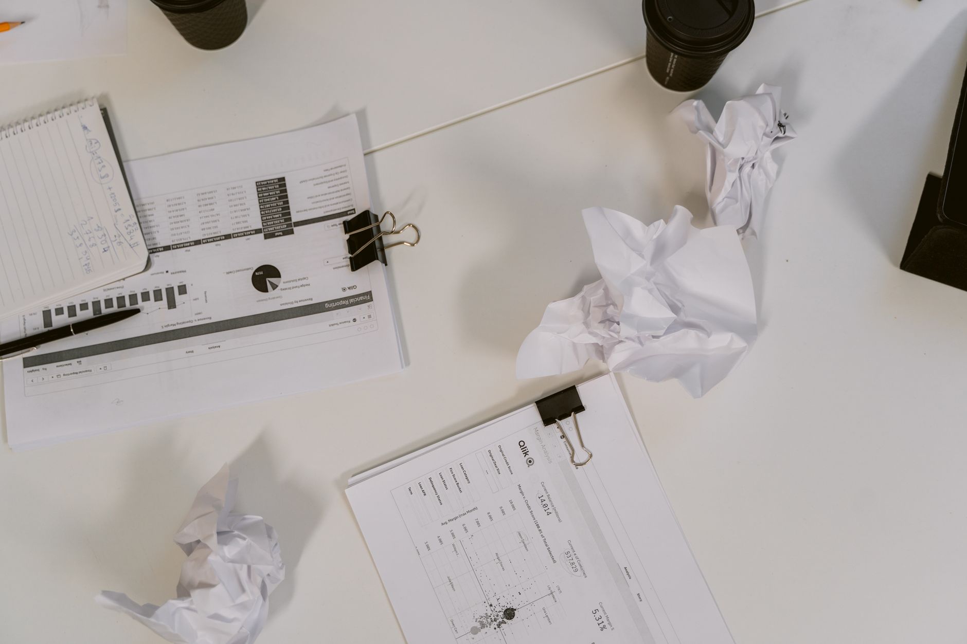 An overhead view of a desk filled with business documents, crumpled paper, and coffee cups.