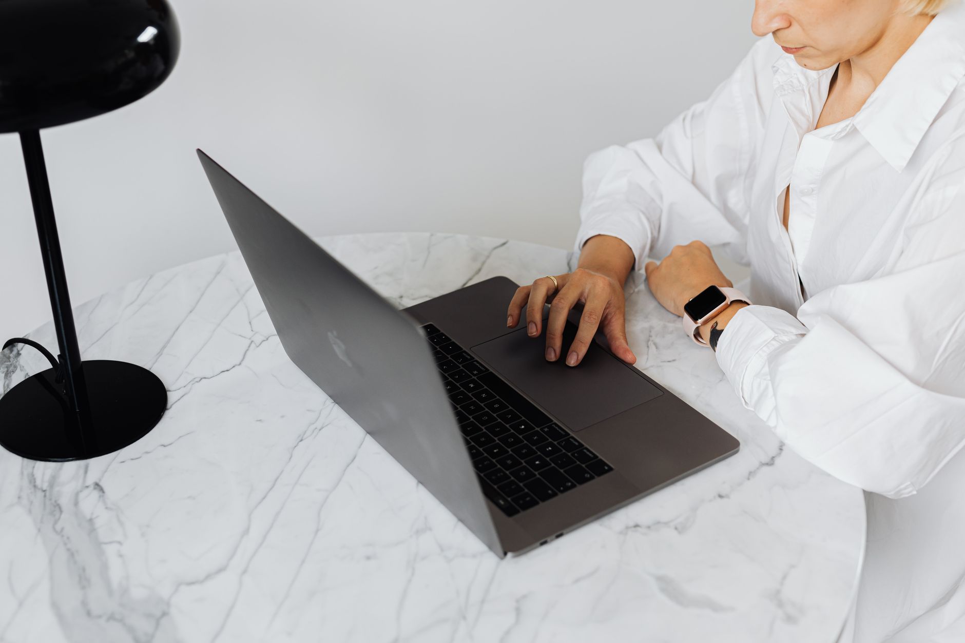 A woman in white shirt working on a laptop at a marble table with elegant desk lamp.
