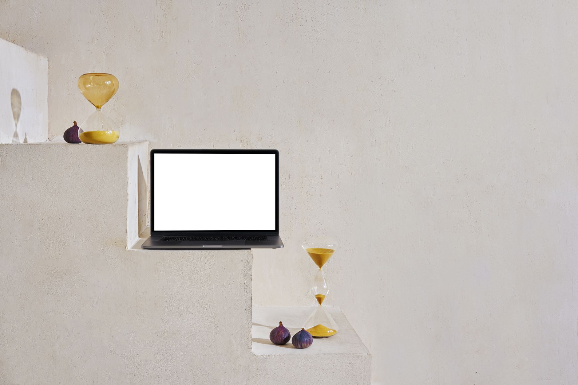 Laptop with white screen placed on white stone steps near sand glasses and small figs near wall in light place